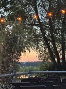 Das Open Air Kino findet in Kreuzlingen im Seeburgpark direkt am See statt. Das Foto wurde von den Sitzen aus gemacht und der Blick auf den See sowie der Sonnenuntergang sind hier abgebildet. Mehrere Bäume bilden wie eine Art Bogen und eröffnen ein Fenster auf den See und den Sonnenuntergang. Am oberen Rand ragt noch eine Lichterkette durch das Foto.