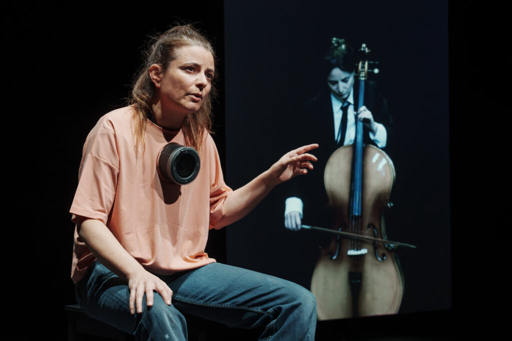 Sarah Hugentobler bei ihrer Performance mit einem tragbaren Lautsprecher am Shirt. Im Hintergrund zeigt eine Videoprojektion sie im Kostüm beim Musizieren.